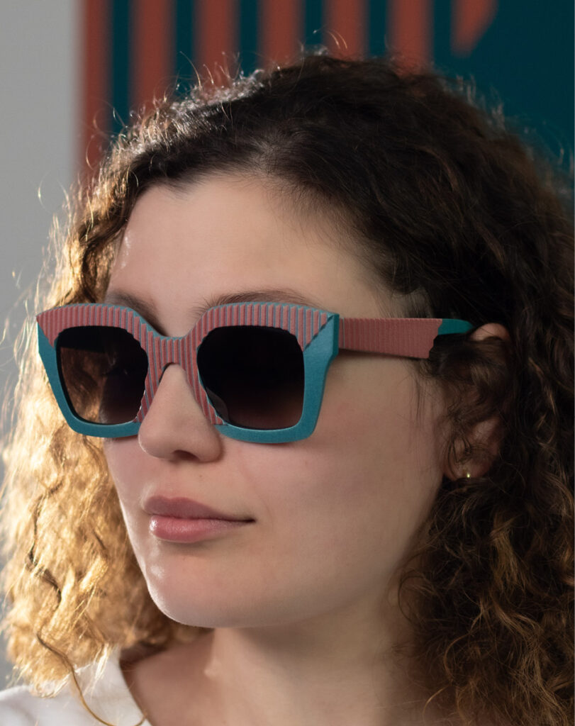 Model wearing AVA Orange Bay sunglasses by CEHO Studio, with teal and coral striped frames, photographed in front of a graphic color-block backdrop.