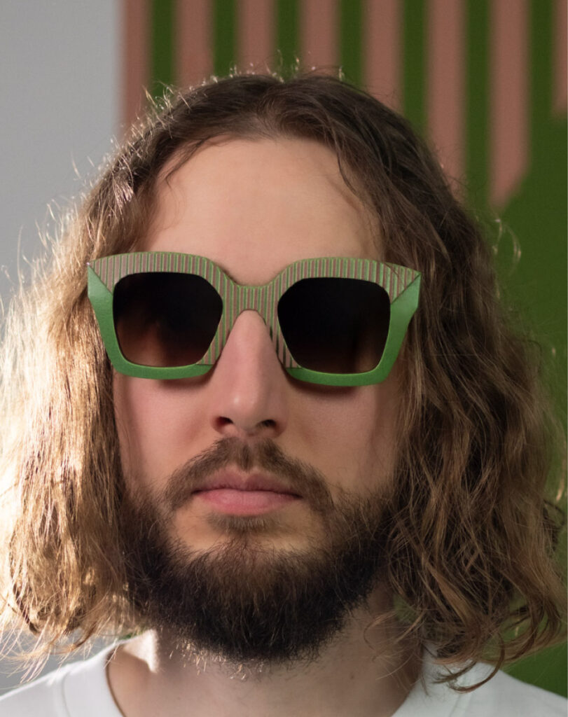 Model wearing AVA Pink Jungle sunglasses by CEHO Studio, featuring green frames with pink-striped detailing against a graphic green and pink backdrop.