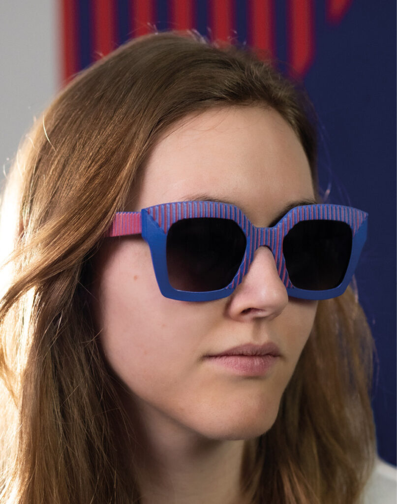 Female model wearing AVA Red Yves sunglasses by CEHO Studio, featuring bold blue frames with red-striped texture, shot against a deep blue and red backdrop.