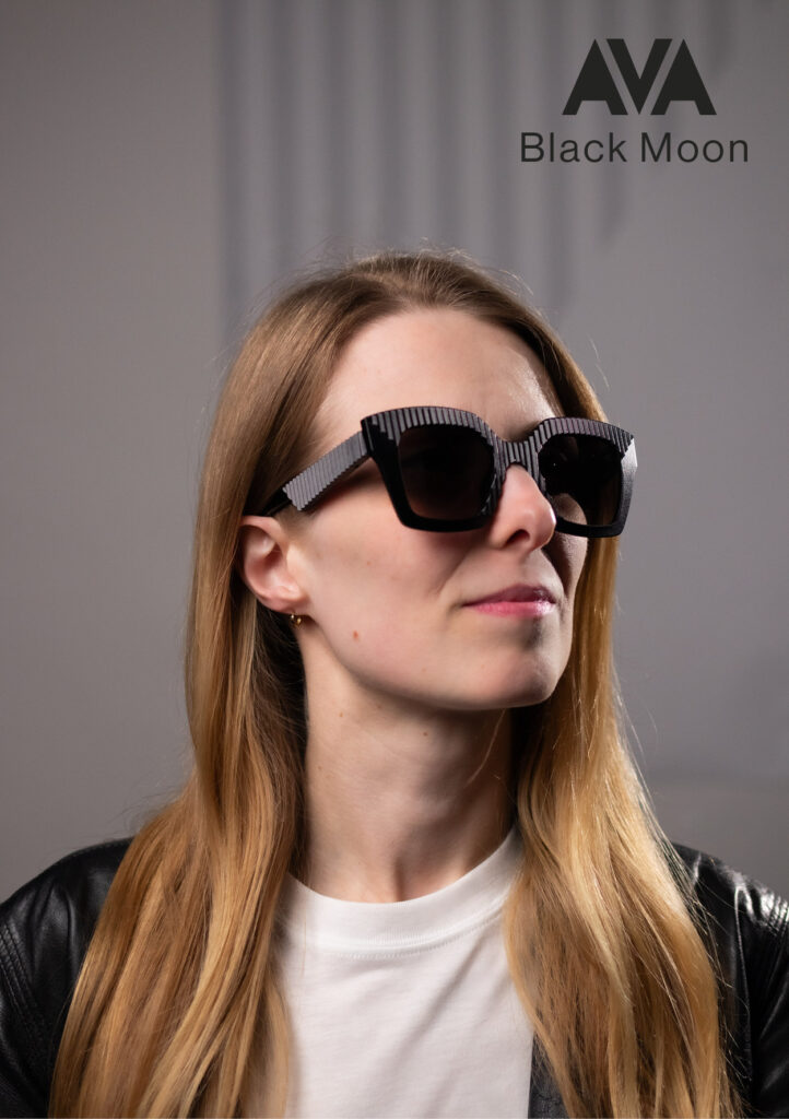 Model wearing AVA Black Moon sunglasses by CEHO Studio, featuring bold black frames with vertical striped detail, set against a moody grey studio background.
