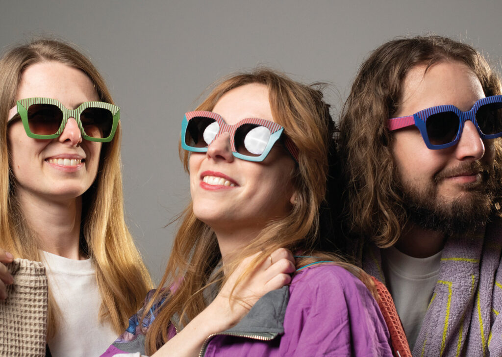 Three models wearing AVA sunglasses by CEHO Studio: Pink Jungle, Orange Bay, and Red Yves variants, smiling together in a playful group portrait against a grey background.